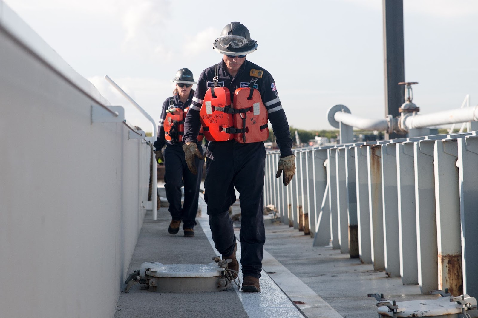 Two people walking along a barge in protective gear and life jackets, as is common with tankerman jobs and as is one of the many key elements of becoming an AccuTrans tankerman.